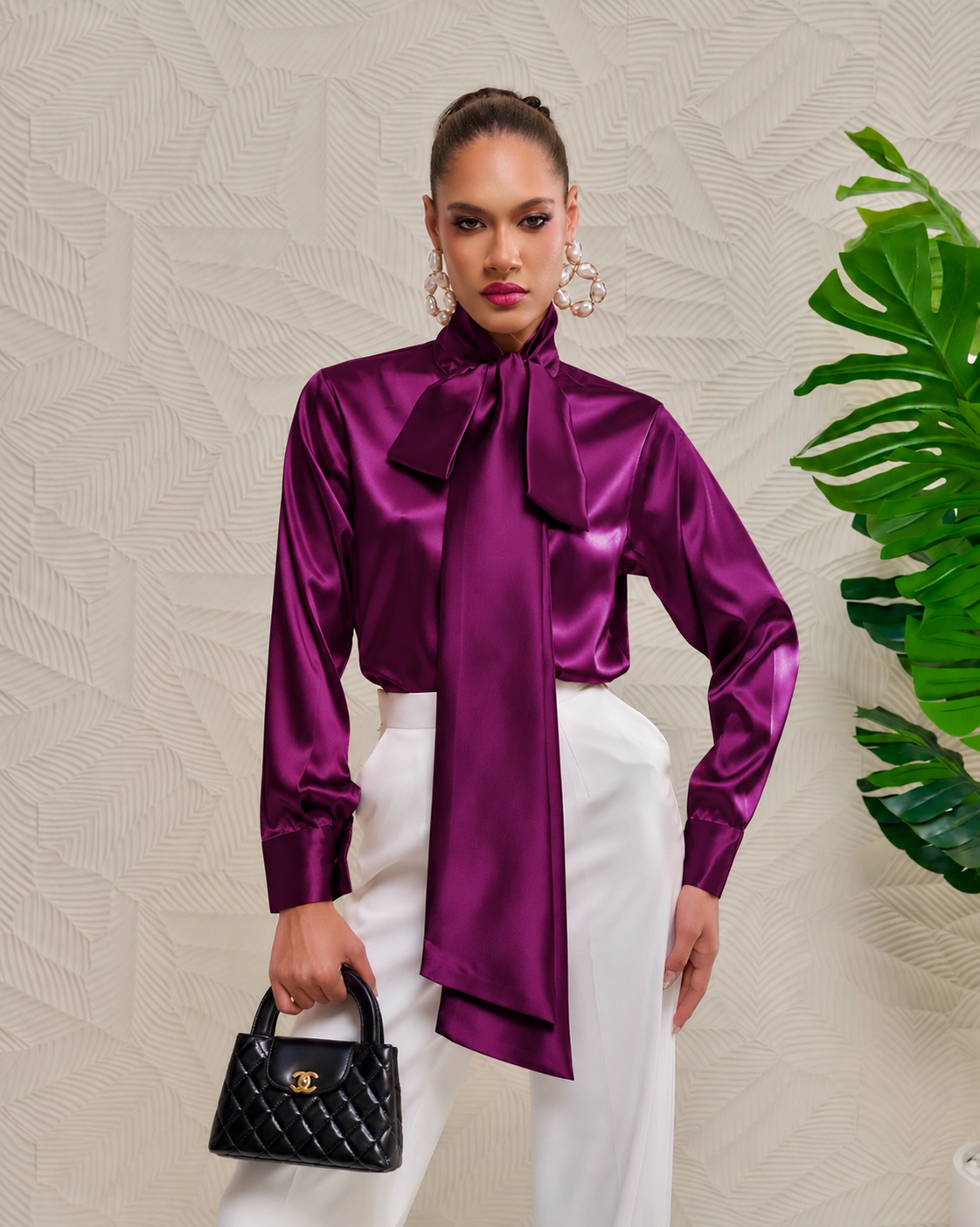 A model wearing a shiny purple satin long-sleeve blouse with a large bow tie-neck detail, paired with white trousers and holding a black quilted handbag, set against a textured beige wall with a green plant leaf on the right.