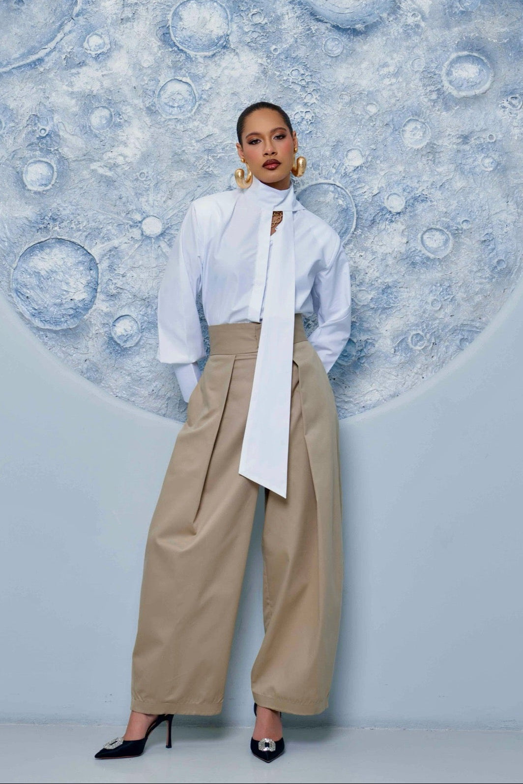 Person wearing a white blouse and beige cargo pants standing in front of a circular wall art with abstract design.