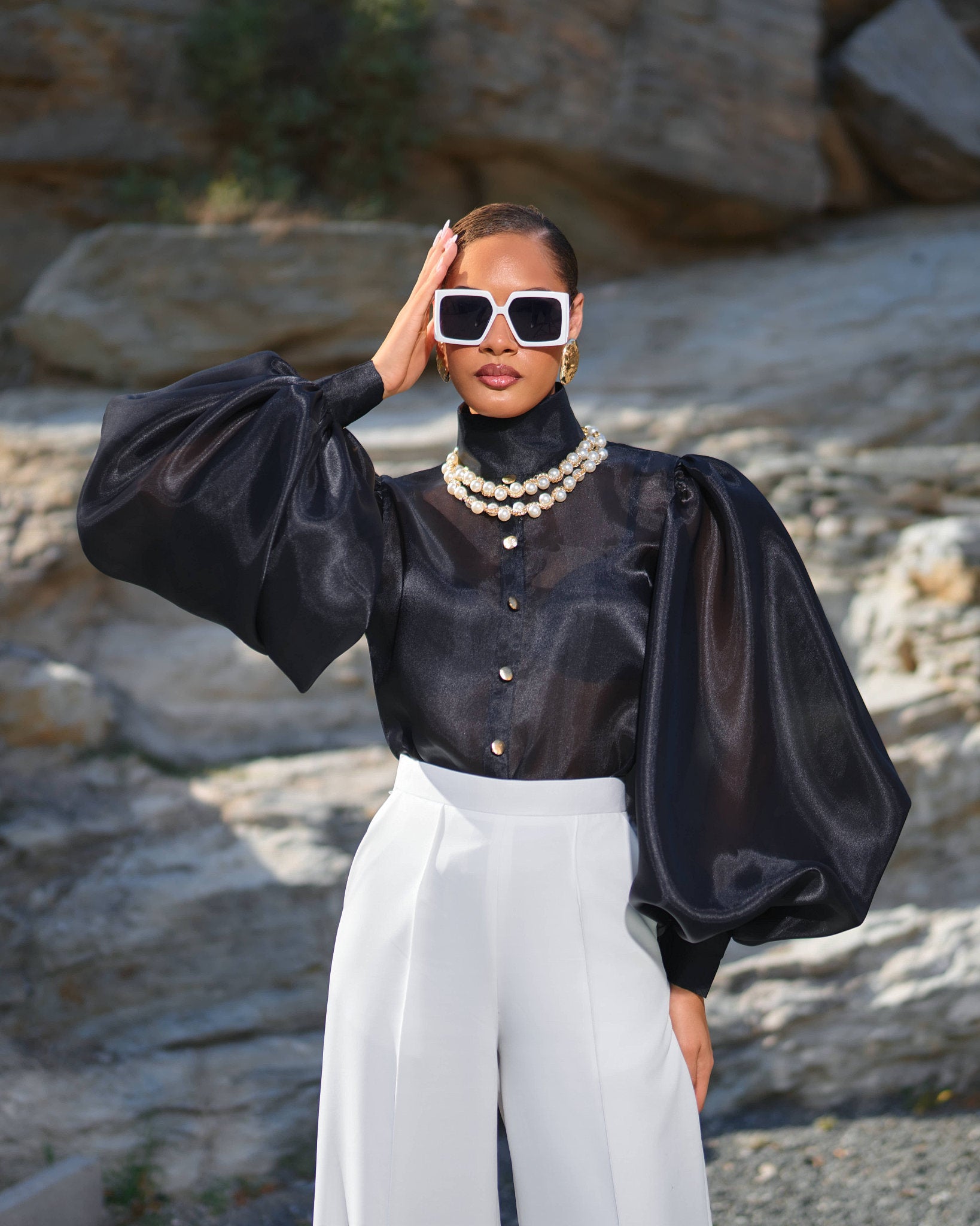A model wearing a sheer black satin blouse with dramatic balloon sleeves, white high-waisted trousers, a double pearl necklace, and white-framed sunglasses, posed outdoors against a rocky background.