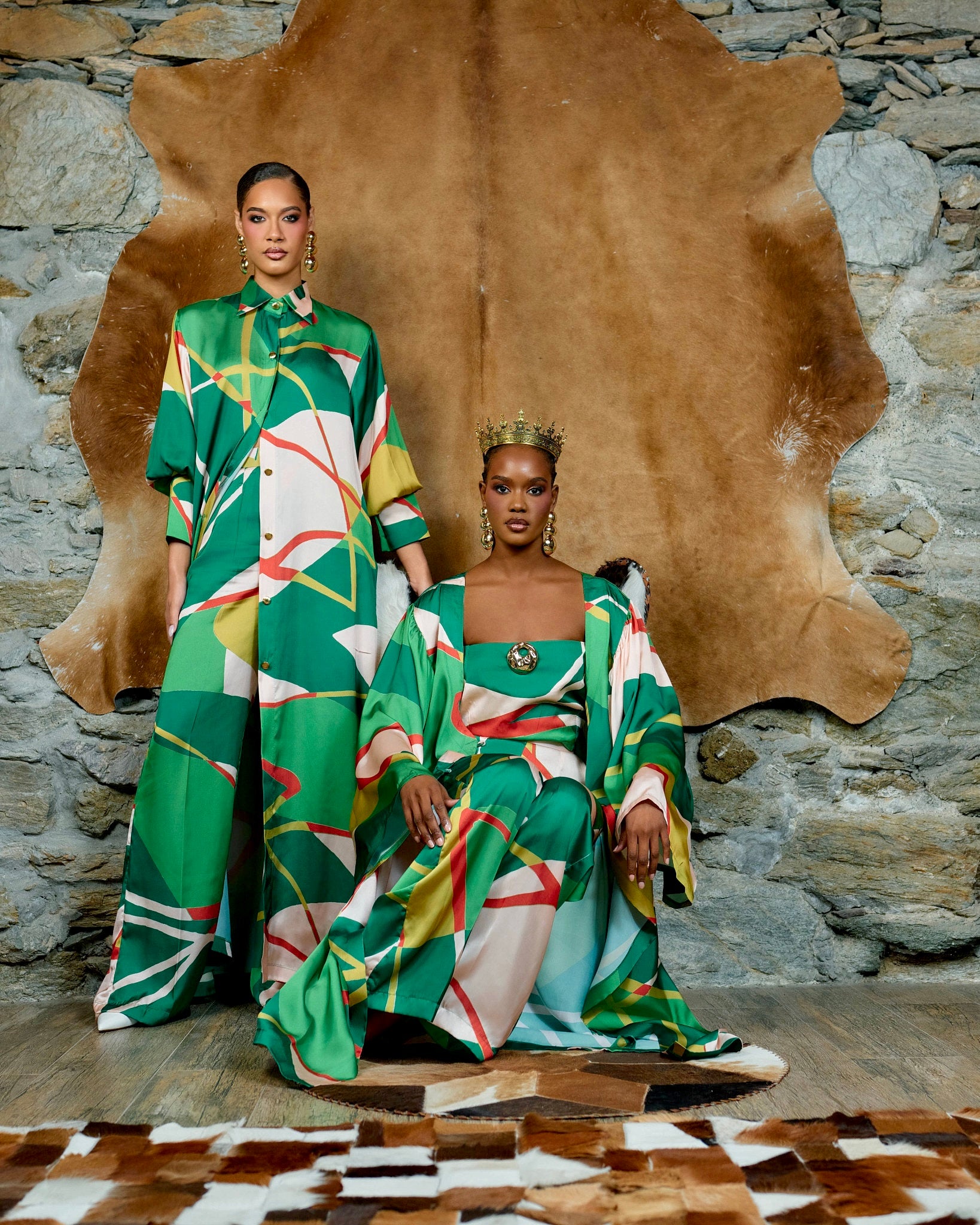 Two models pose in matching green, white, yellow, and red geometric print gowns and caftans in front of a rustic stone wall with a large brown hide backdrop.