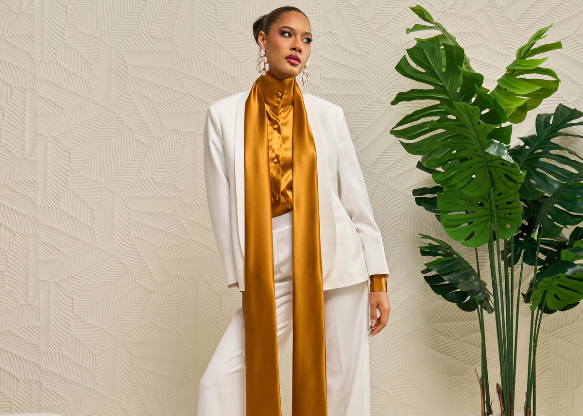 A model stands in a studio setting wearing a white tailored suit with wide-leg trousers and a high-neck silk top in a rich mustard-gold color, with a long silk  detail draping from the neck down. She is posed next to a large potted green plant against a textured beige wall.