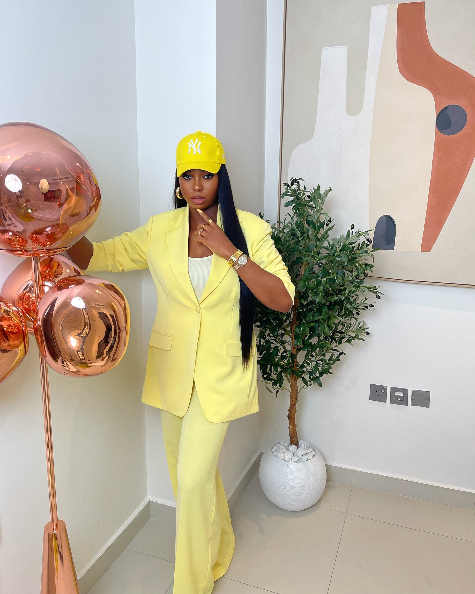 Butter yellow power suit set featuring a structured blazer and straight-leg trousers, styled with gold accessories and a yellow baseball cap.