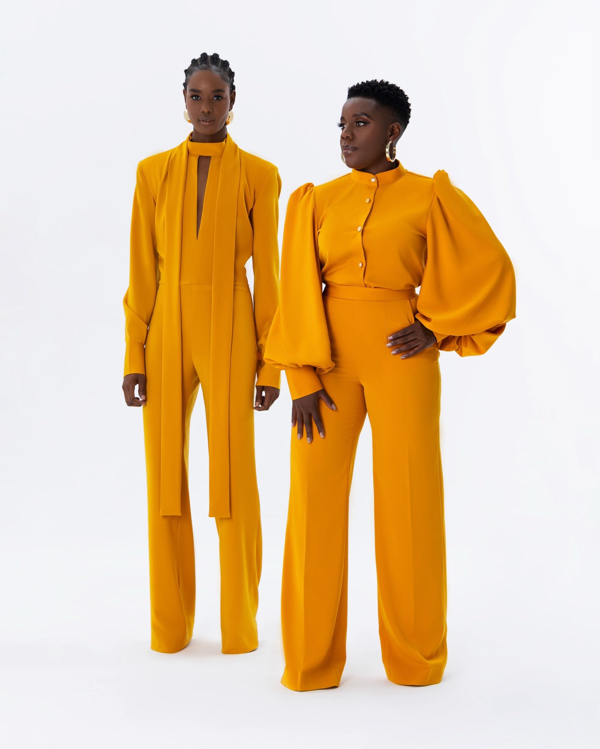 Two models wearing elegant and timeless mustard yellow outfits. One is wearing a stylish mustard yellow jumpsuit with neck tie details and deep v cut, while the other model is wearing a chic monotone matching coord set with balloon sleeve top paired with a high-waist straight pants.