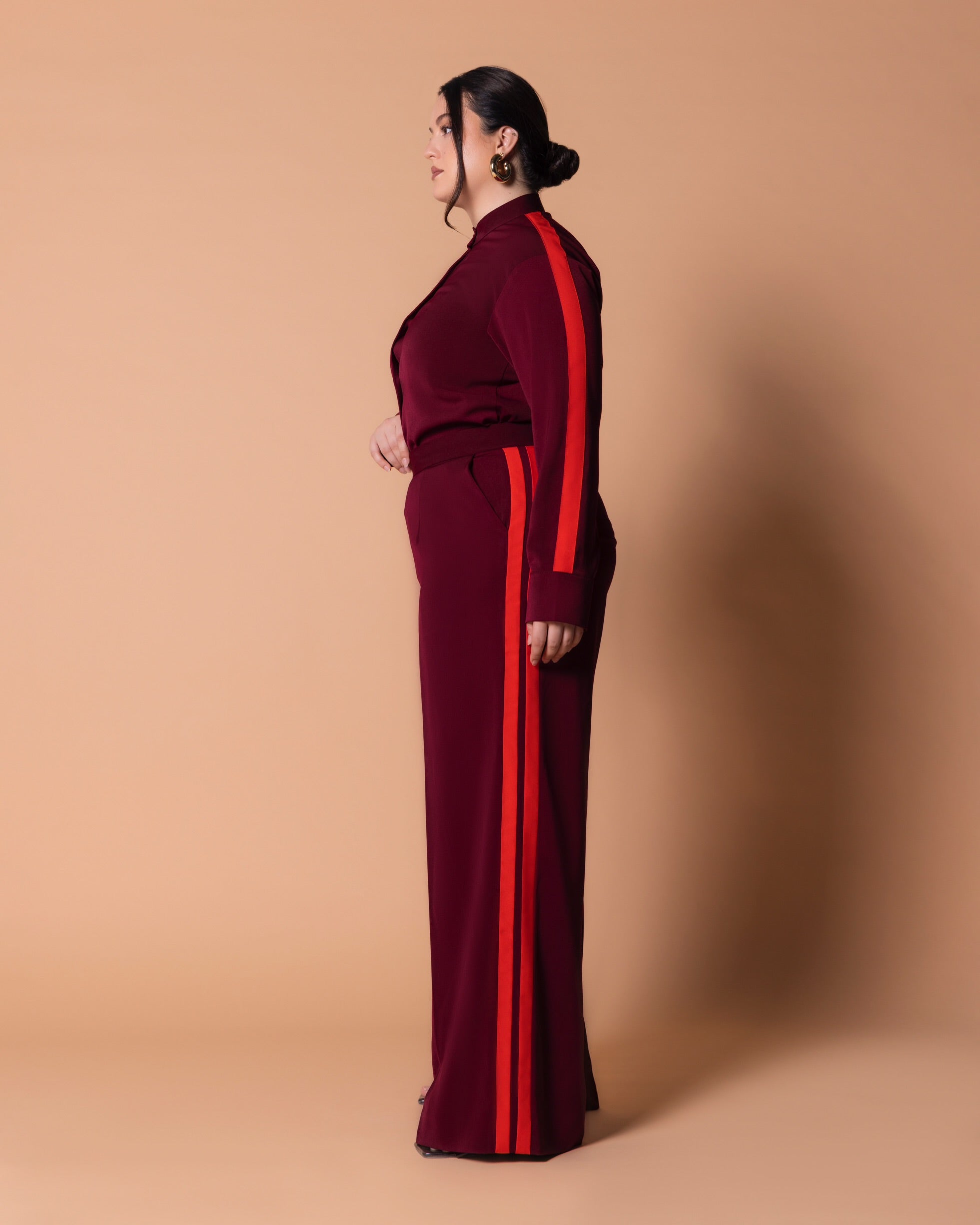 Model in Maison Set wearing burgundy bishop-collar button shirt with orange sleeve stripe and matching high-waist straight-leg pants with double side stripes, side view.