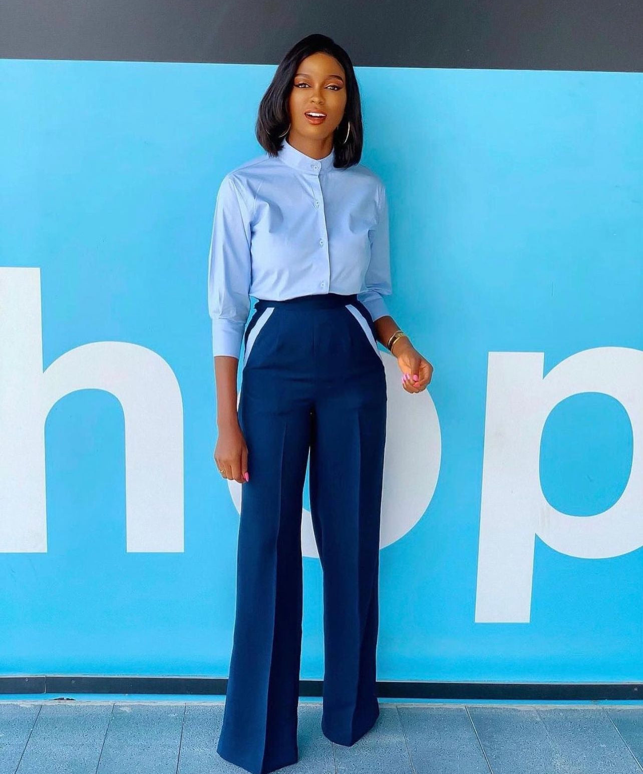 A woman standing in front of a blue wall, wearing a light blue button-down shirt and navy high-waist palazzo pants with side zip and metal hook closure, with sky blue stripes at the pockets.