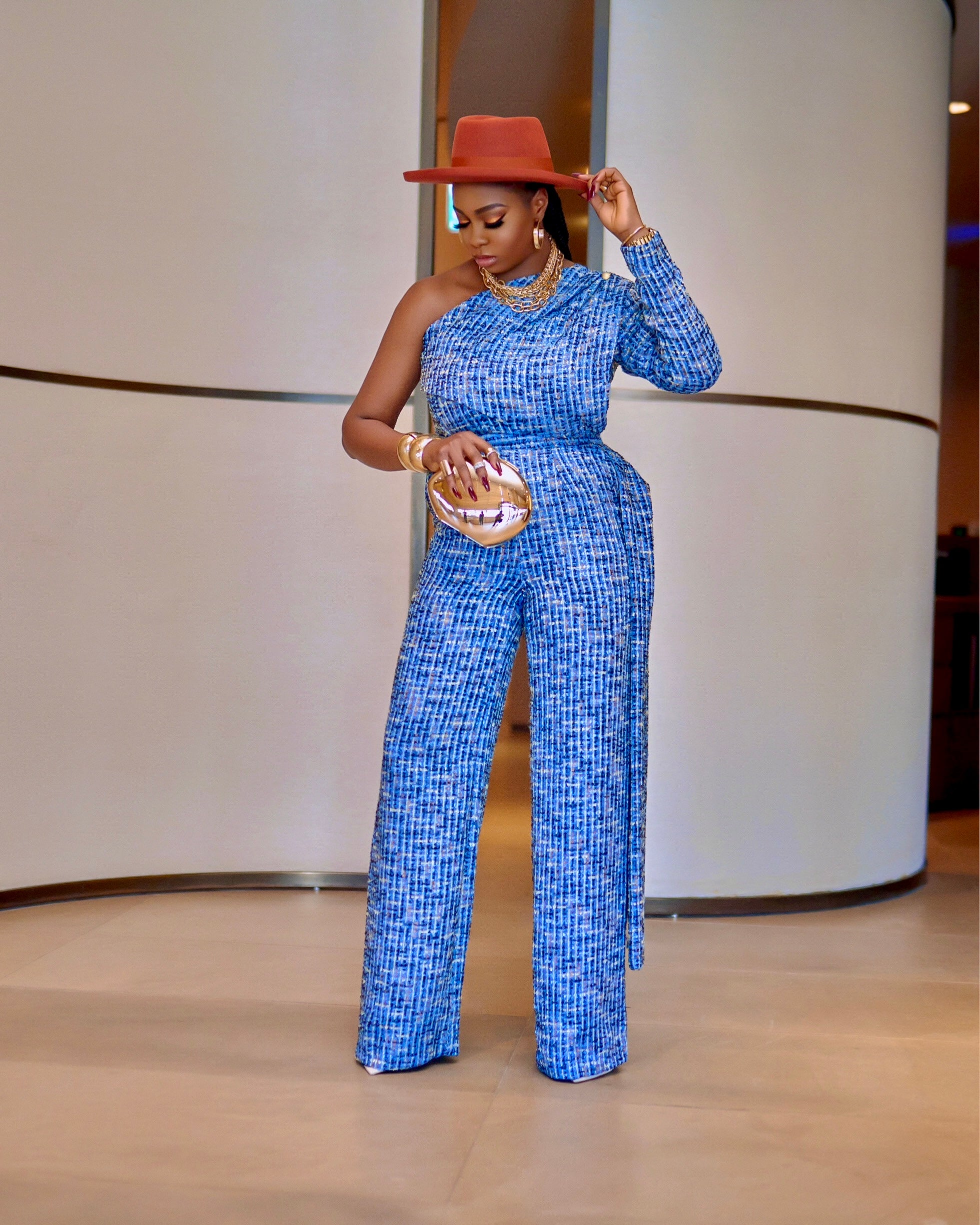 Model wearing Neka blue textured tweed one-shoulder set with waist drape detail and high-waist straight-leg pants, styled with a rust hat and metallic clutch.