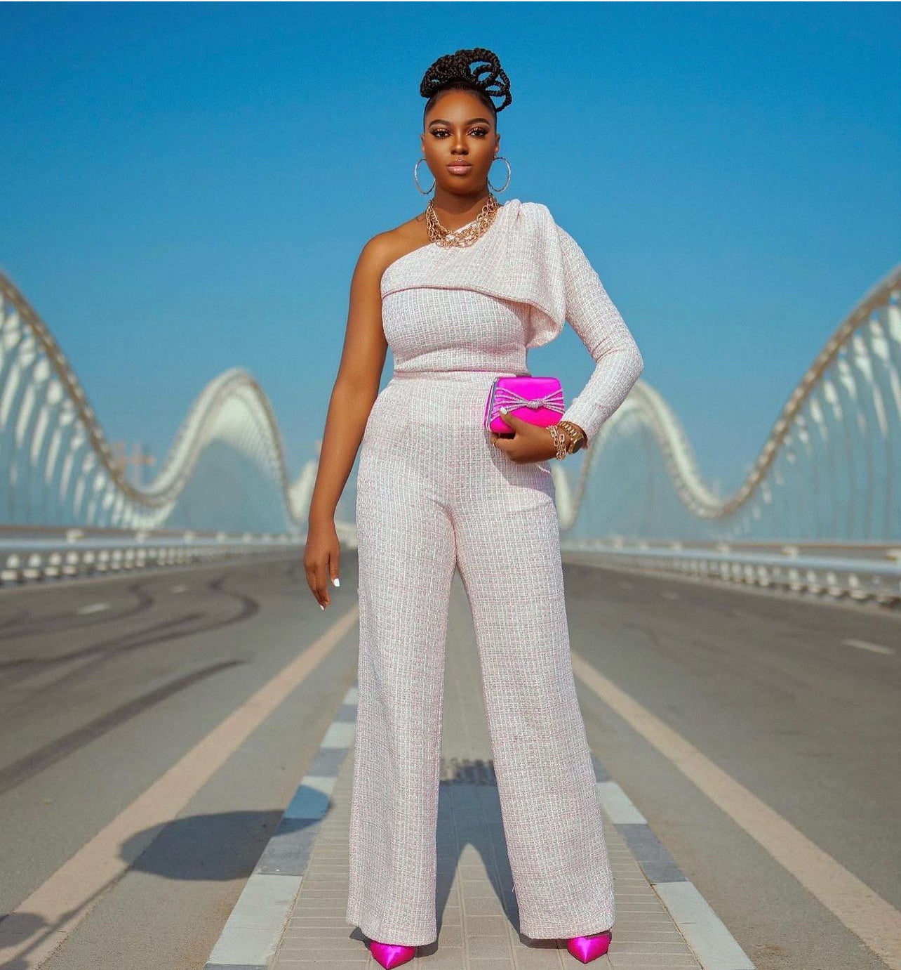A model wearing a luxury long-sleeved pink tweed top with off-shoulder, bow-like detailing on the front and a gold button on the shoulder. It's paired with high waist pants with pockets and a side zipper. Styled with hot pink Jimmy Choo clutch and shoes. 

The model is standing in the middle of the Maydan bridge in the city of Dubai.