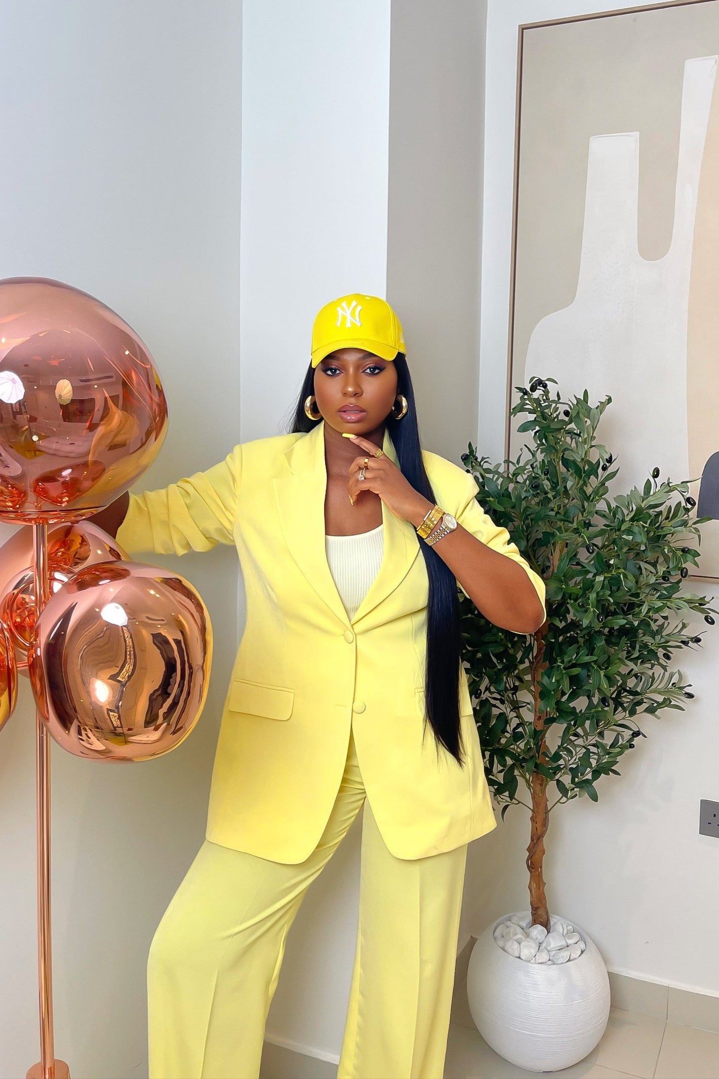 Model wearing Noura butter yellow suit set with tailored blazer and a high-waist, straight-leg pants, styled with a yellow cap and white top indoors.