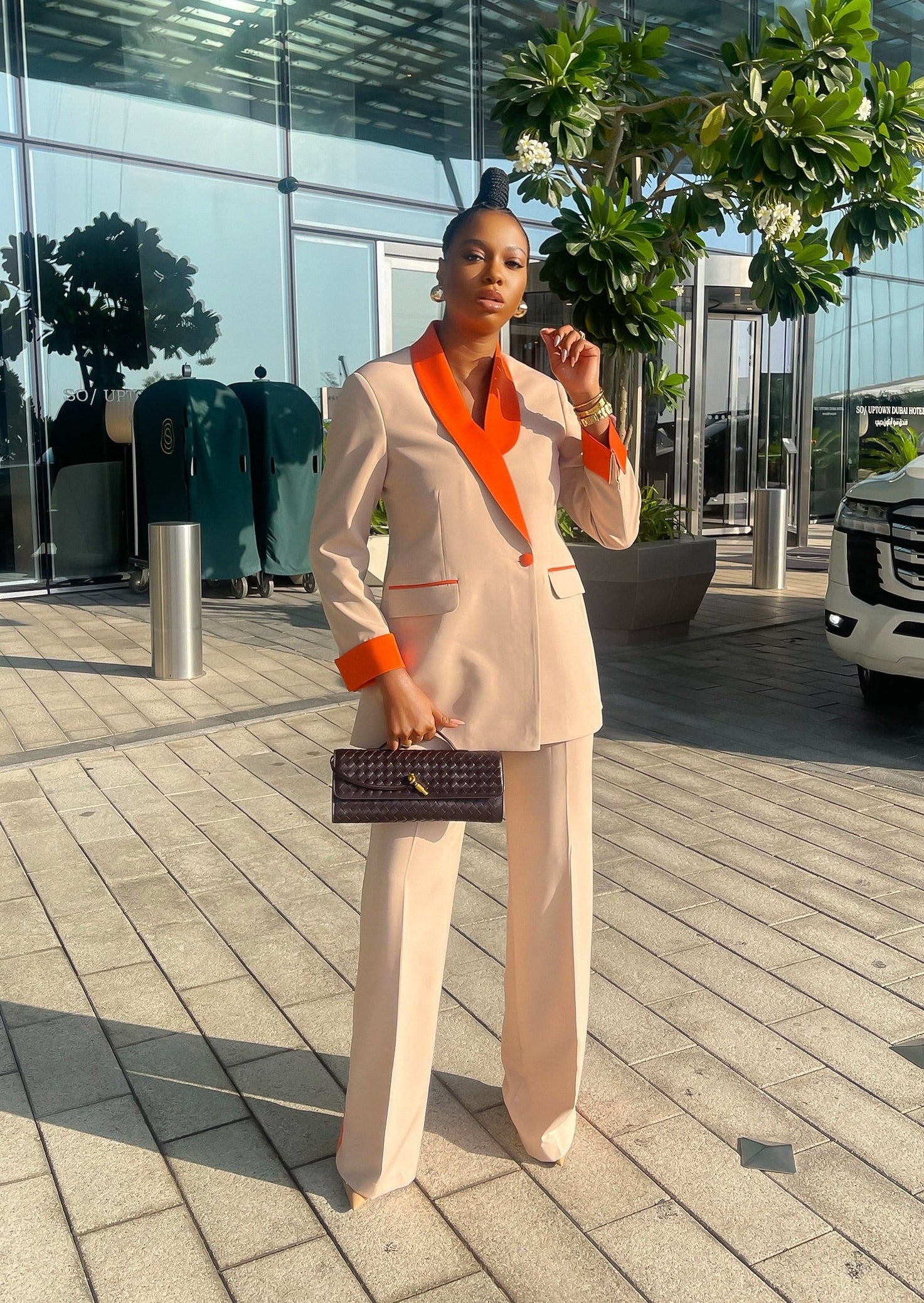 A stylish woman in a nude and orange double-breasted suit with orange turn-up cuffs and bold side-striped trousers, standing confidently in an urban setting.