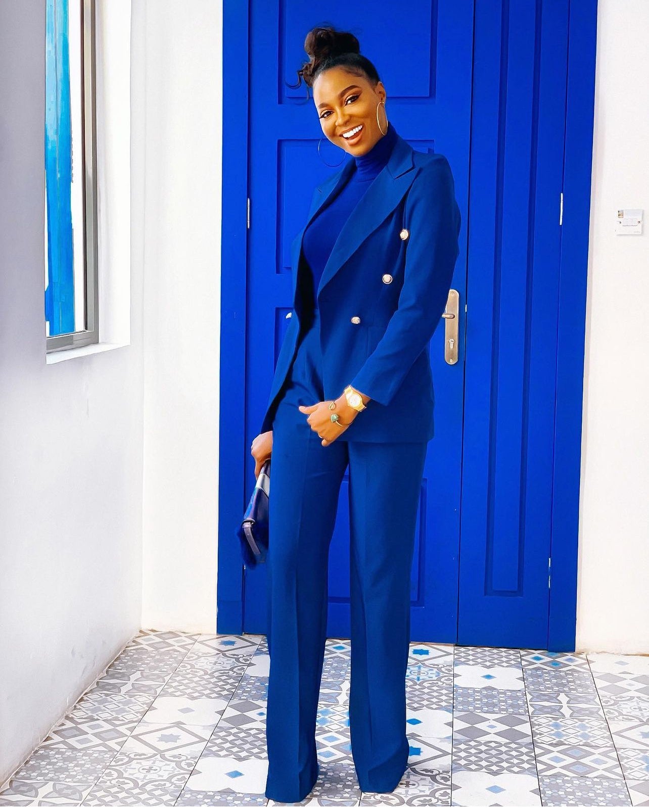 A model standing in front of a blue door wearing a double-breasted blue blazer with lapel collar and silver buttons, paired with matching high-waisted pants with side zip and metal hook closure.