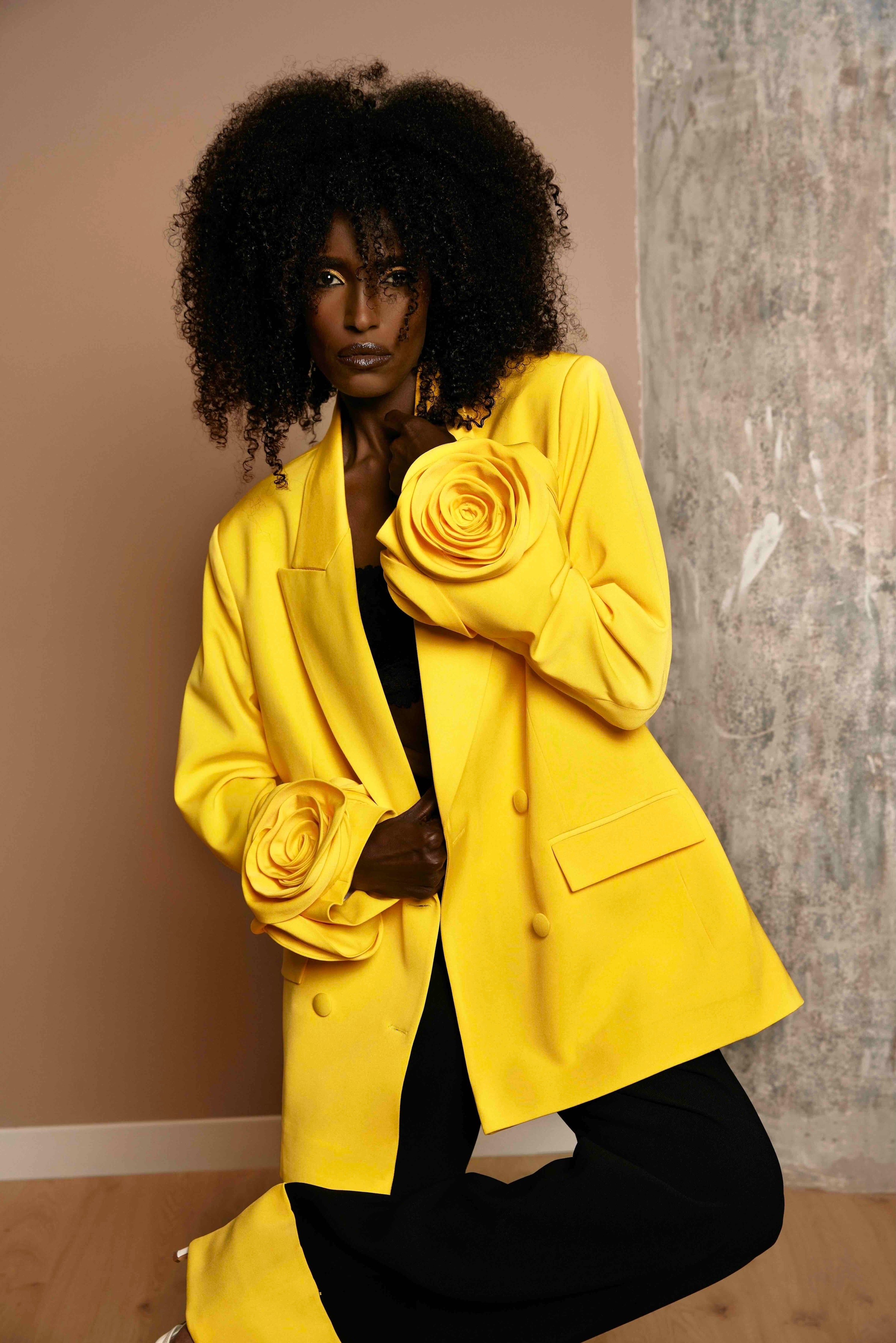 A dark skin model wearing a sunny yellow oversized blazer with floral applique details on both sleeves, paired with black trousers with yellow turn up and floral appliques.