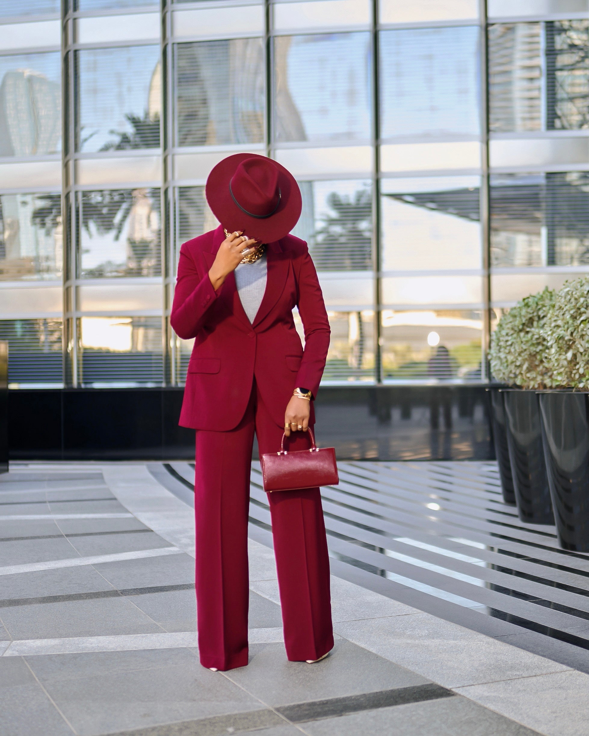 Model is wearing a structured Olarsgrace  burgundy suit set with single-breasted blazer (covered button) and matching straight-leg trousers, styled with hat and mini bag for a luxe monotone look.