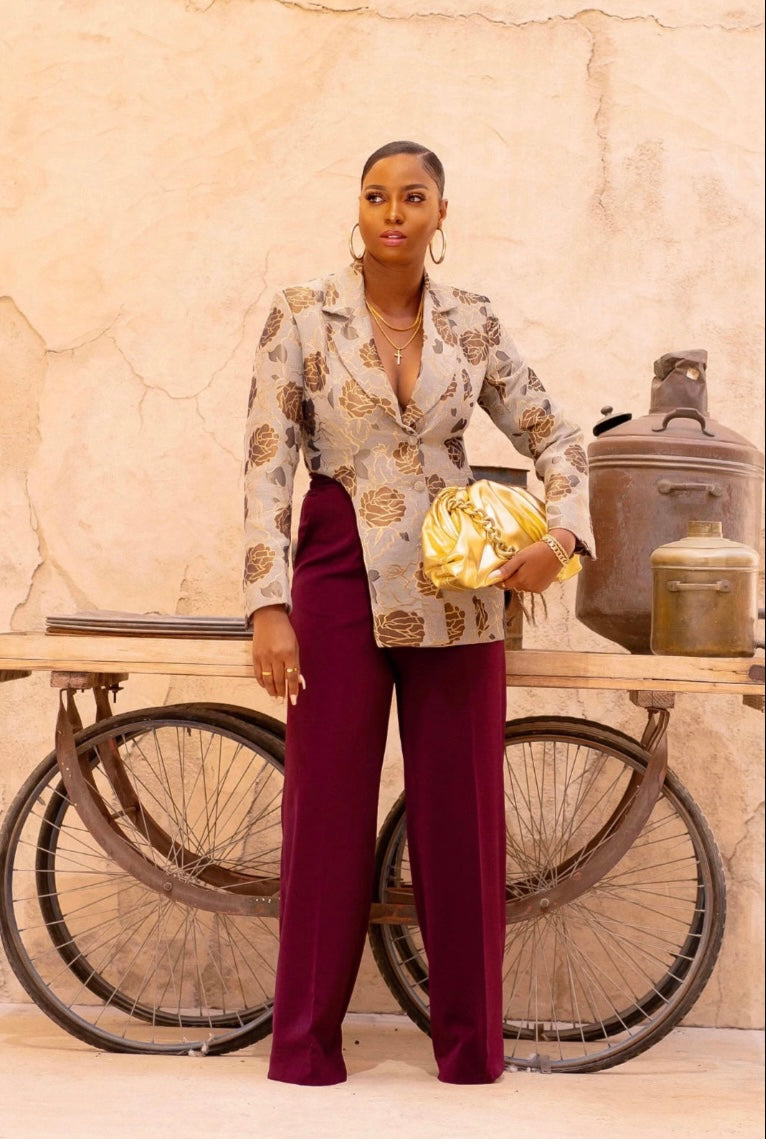 Woman wearing a statement blazer in luxurious grey and gold brocade, posing in front of a rustic backdrop with a vintage bicycle.