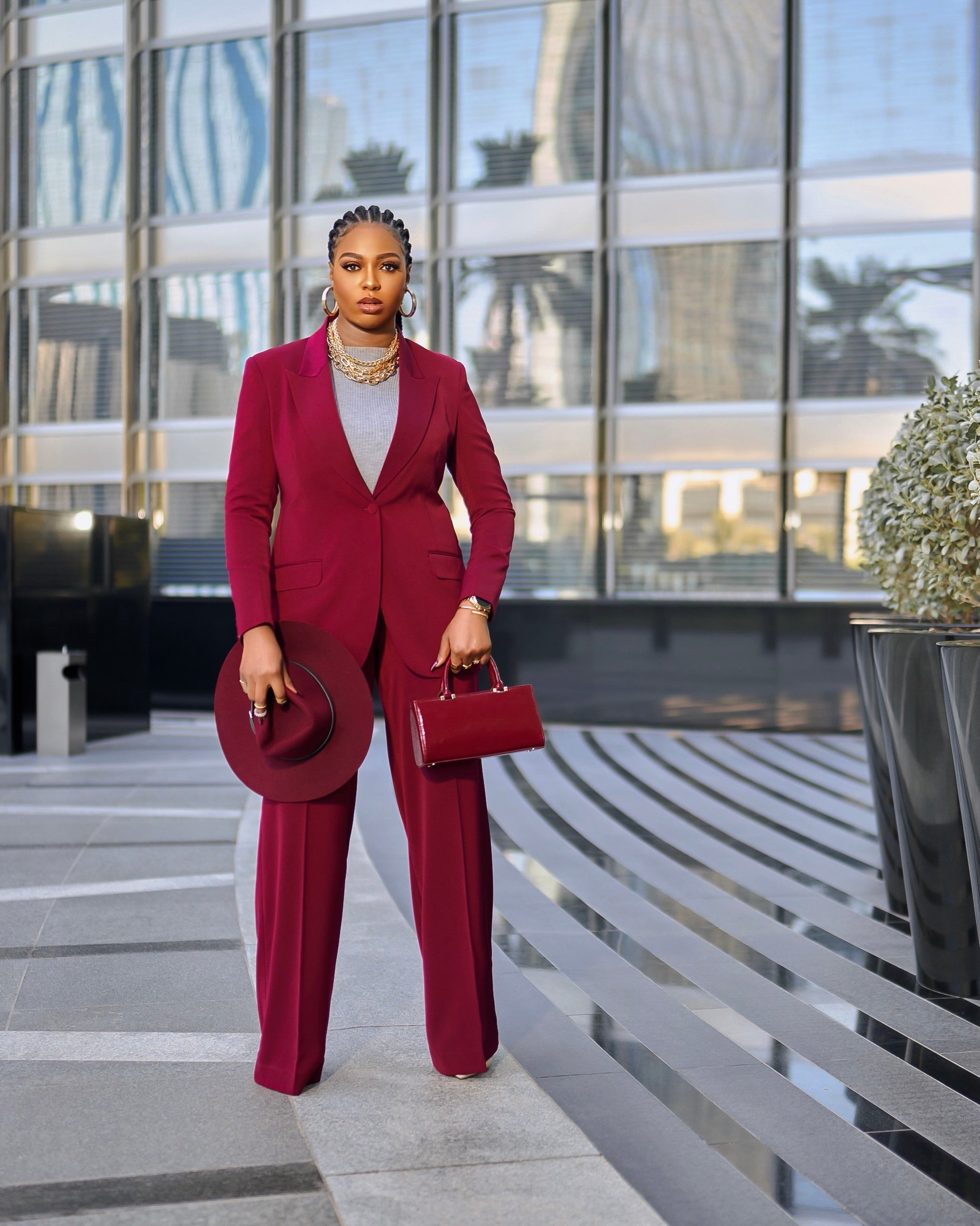 Model is wearing a structured Olarsgrace  burgundy suit set with single-breasted blazer (covered button) and matching straight-leg trousers, styled with hat and mini bag for a luxe monotone look. Photographed outdoors in Dubai, a modern city setting.
