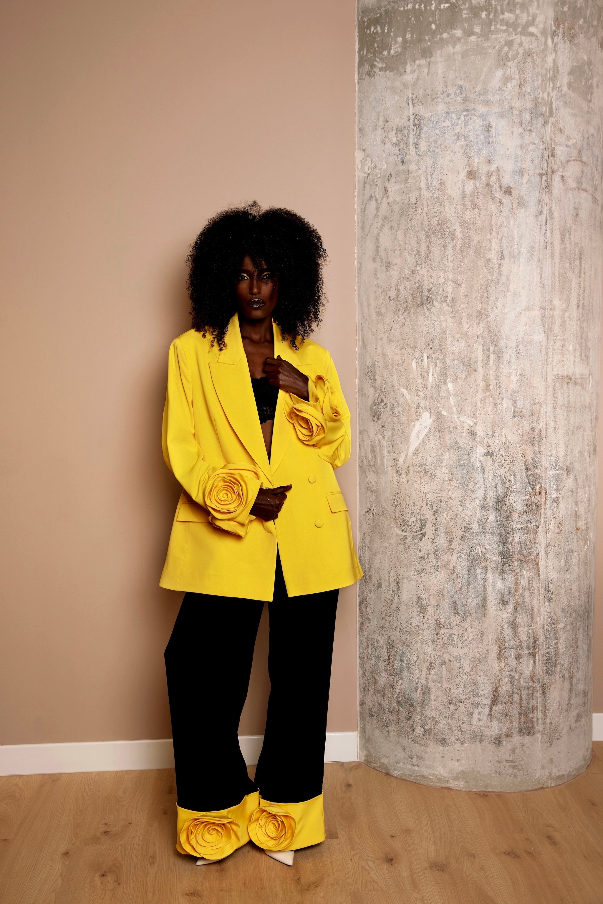Dark-skinned model  in a sunny yellow oversized blazer with floral appliqués on the sleeves, paired with black trousers featuring yellow turn-ups and matching floral details.