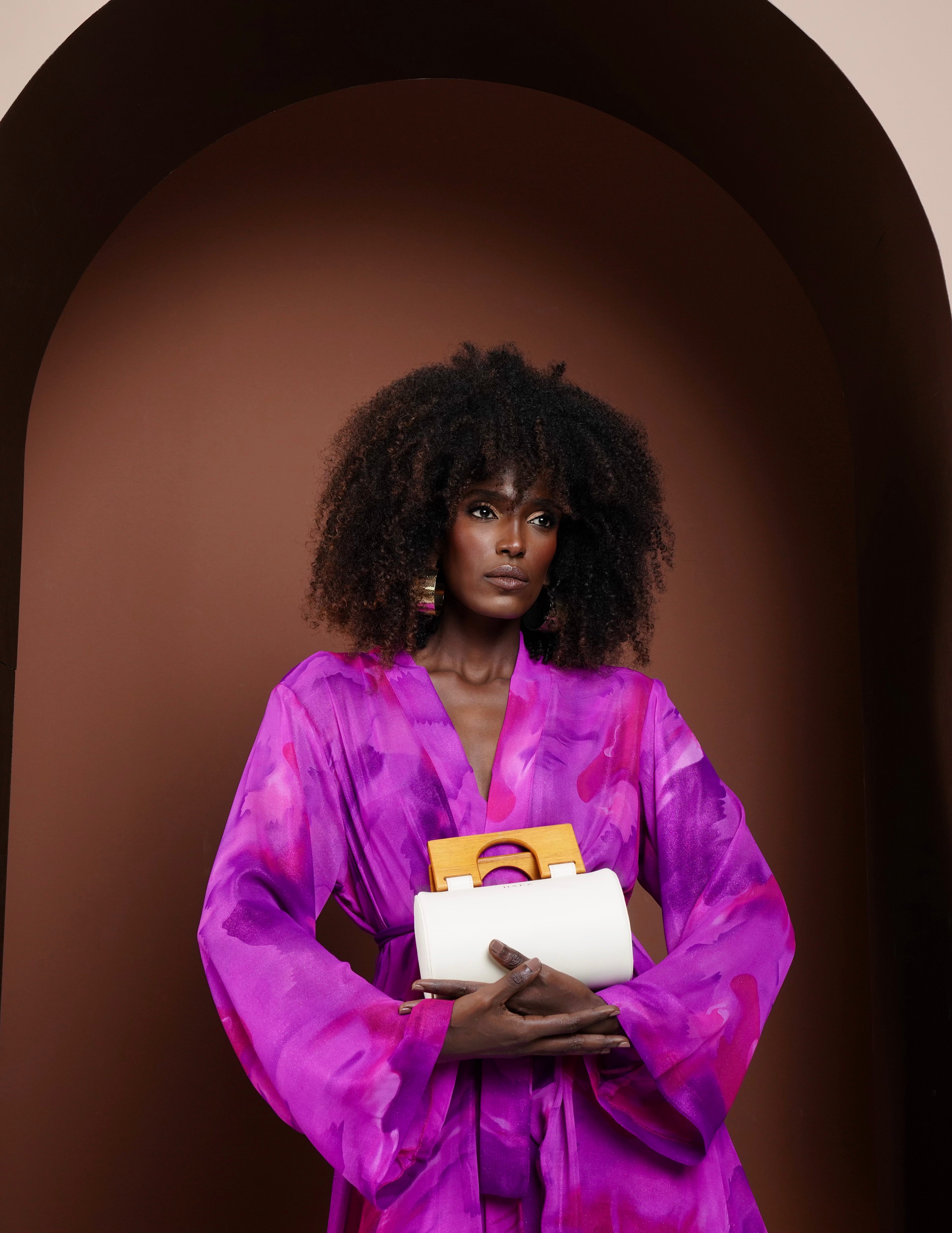 Woman in a vibrant purple kimono outfit holding a white clutch against a brown background