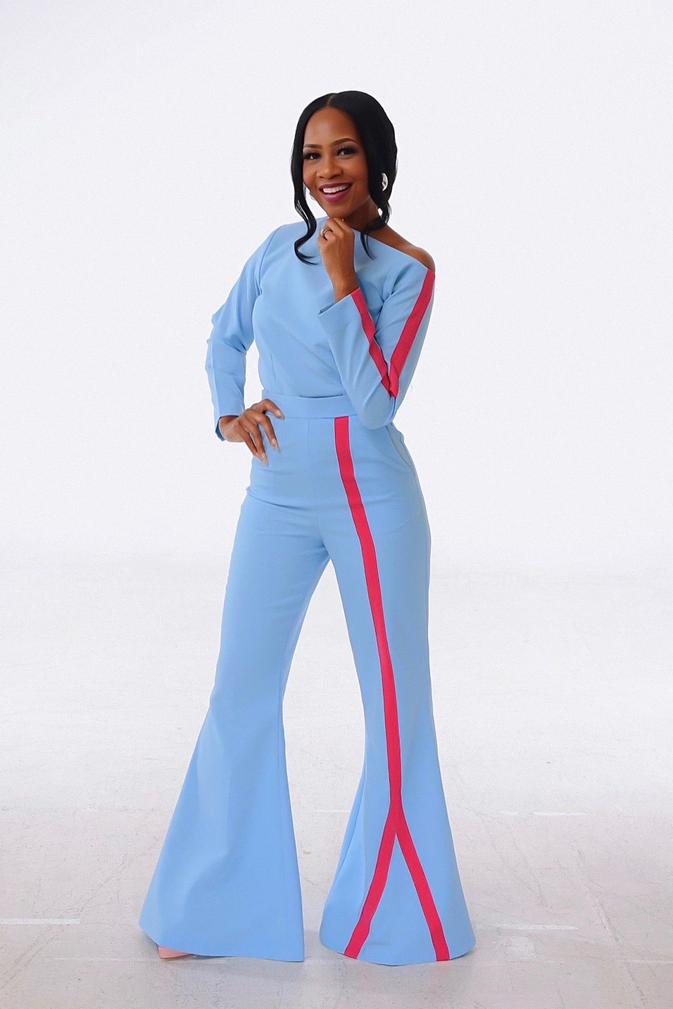 A woman wearing a blue pant set featuring an off-shoulder top, and bell bottom high-waist pants with a bold colour stripe on one leg. She is posing against a white background.