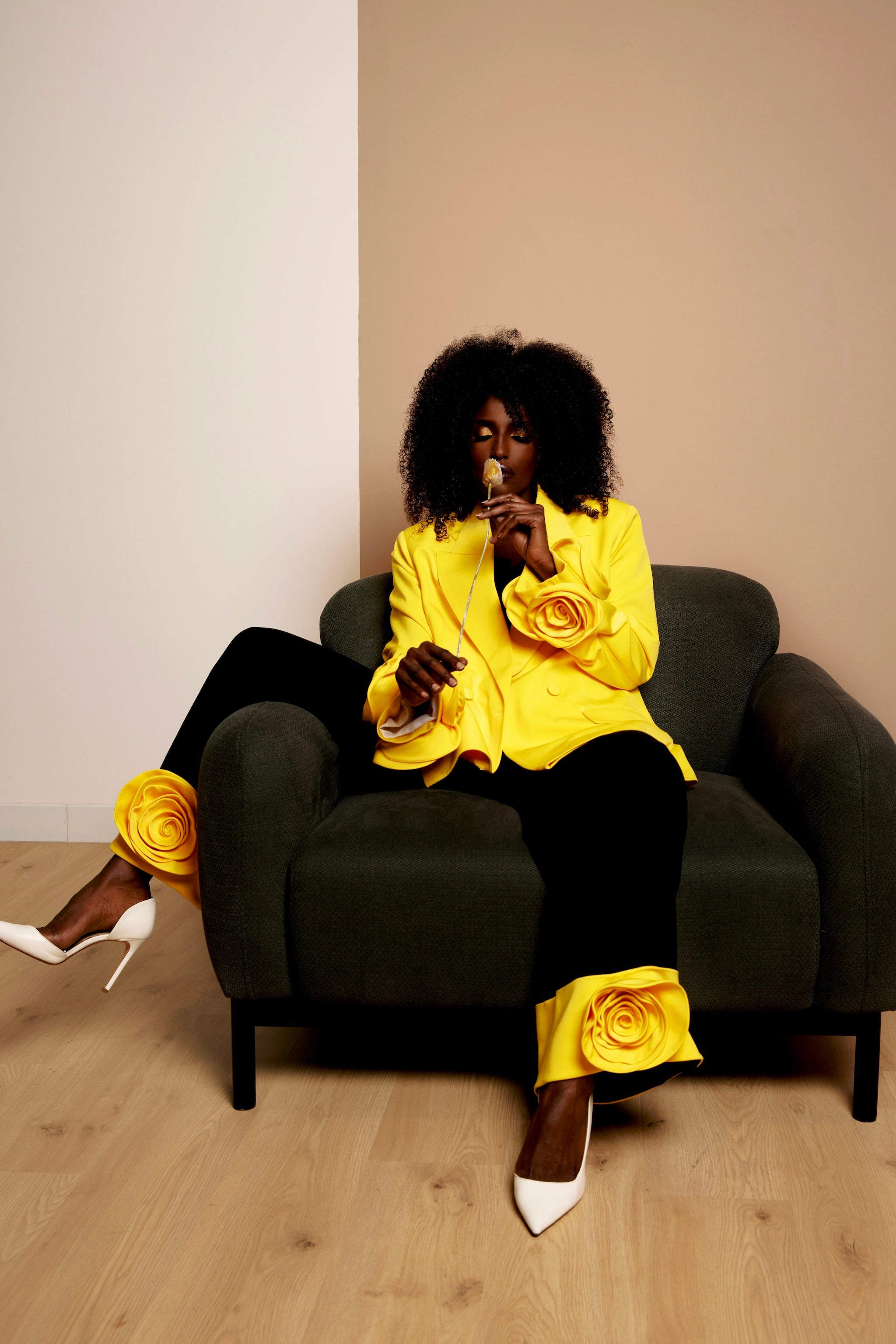 Dark-skinned model seated in a sunny yellow oversized blazer with floral appliqués on the sleeves, paired with black trousers featuring yellow turn-ups and matching floral details.