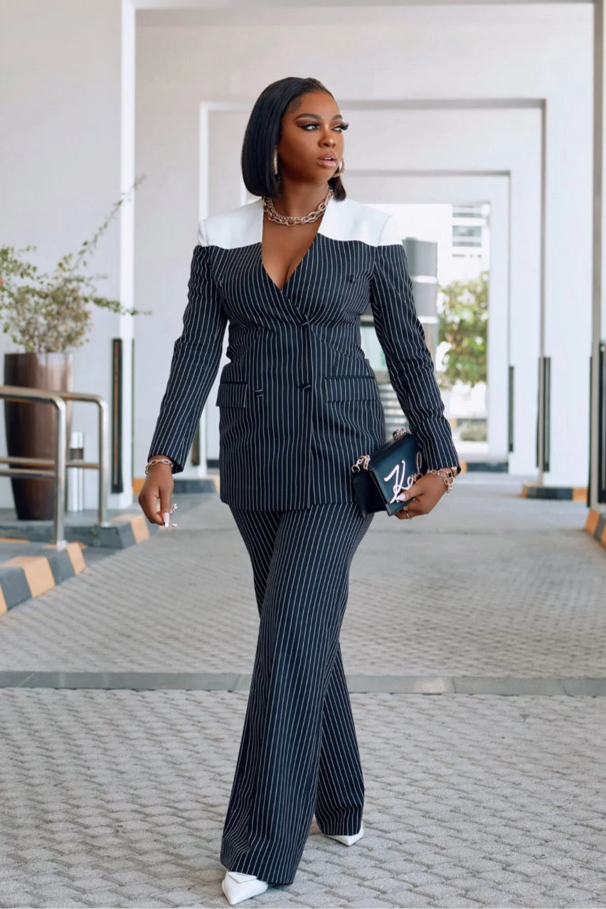 Model walking confidently in a fitted monochrome pinstripe blazer and pants suit with white accents, styled with white heels and bold accessories.