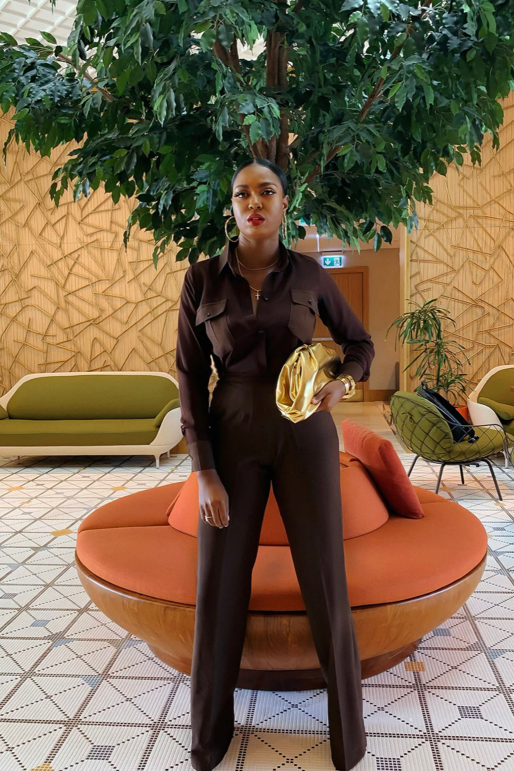 A woman standing in a contemporary lobby with a large tree in the background, wearing an elegant brown long sleeved chiffon shirt with button closure and high waist pants with pockets, holding a chic gold bag and with a side zipper.