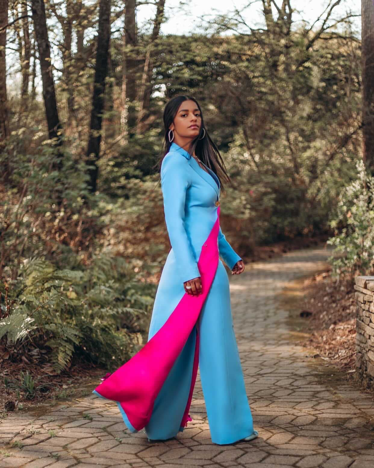 Model is wearing a full-length blazer and pant set in sky blue with pink inner lining and floral beaded detailing, worn by model in a forest backdrop.