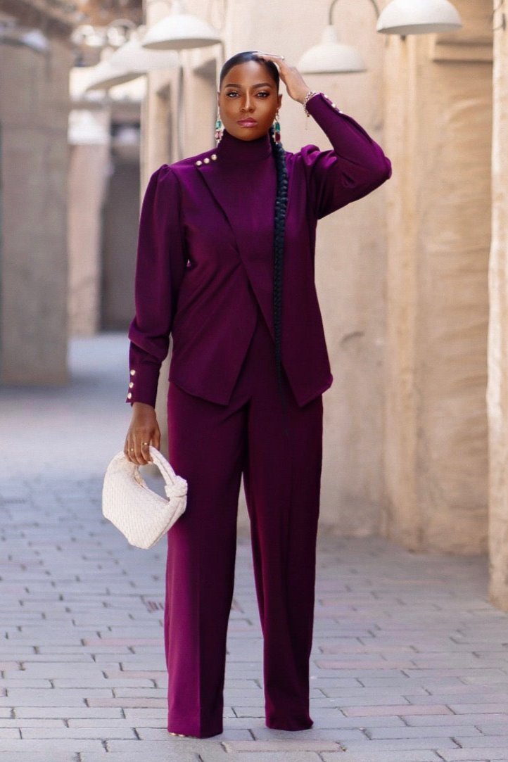 Model wearing the Bako Set in deep purple coord set with an exaggerated turtleneck top, gold button accents on the shoulder and cuffs, and high-waist straight-leg pants, styled with a white bottega handbag.