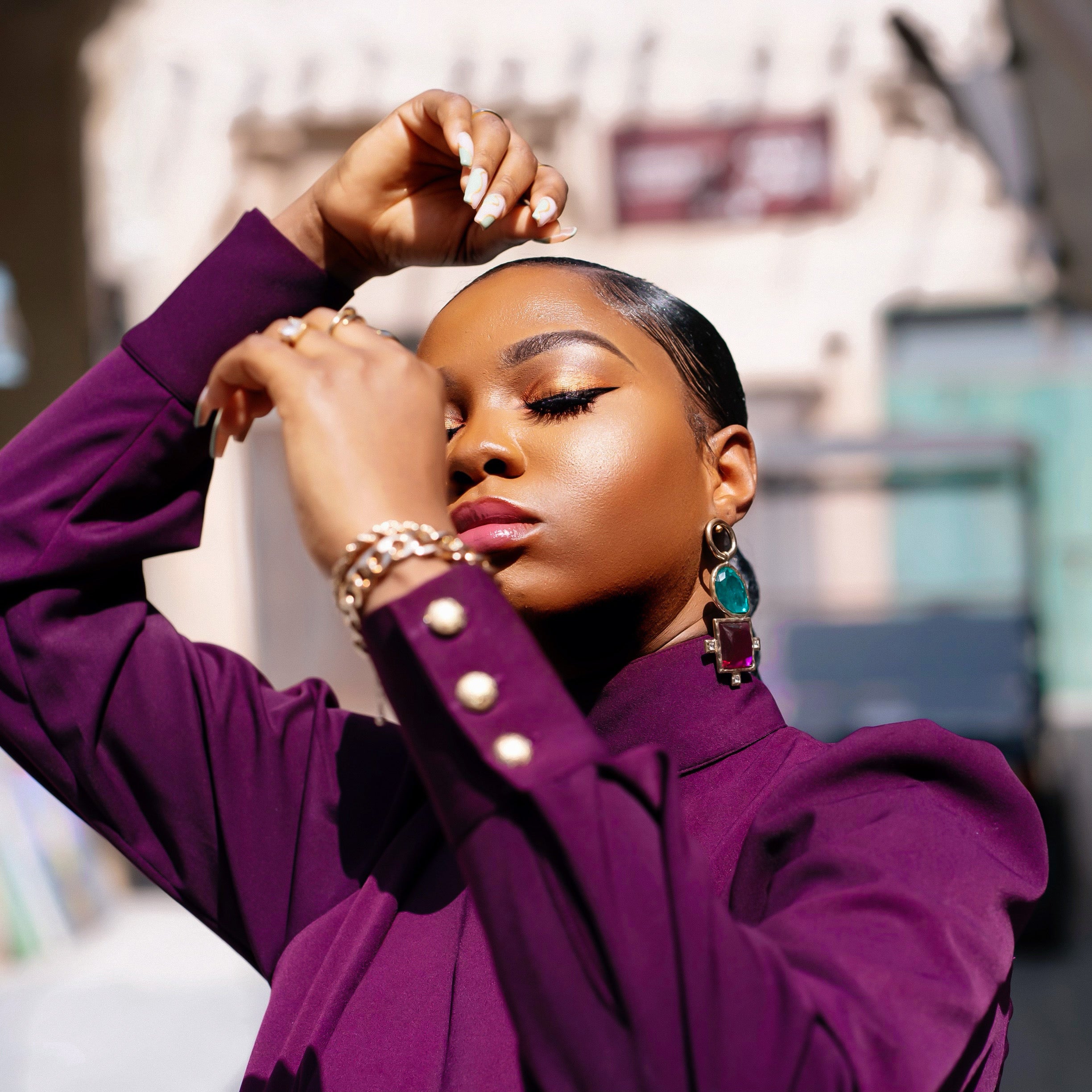Close-up of model in the Olarsgrace Bako Set. A purple aubergine high-neck top highlighting gold accents on the shoulder and cuffs with statement earrings and soft glam makeup.