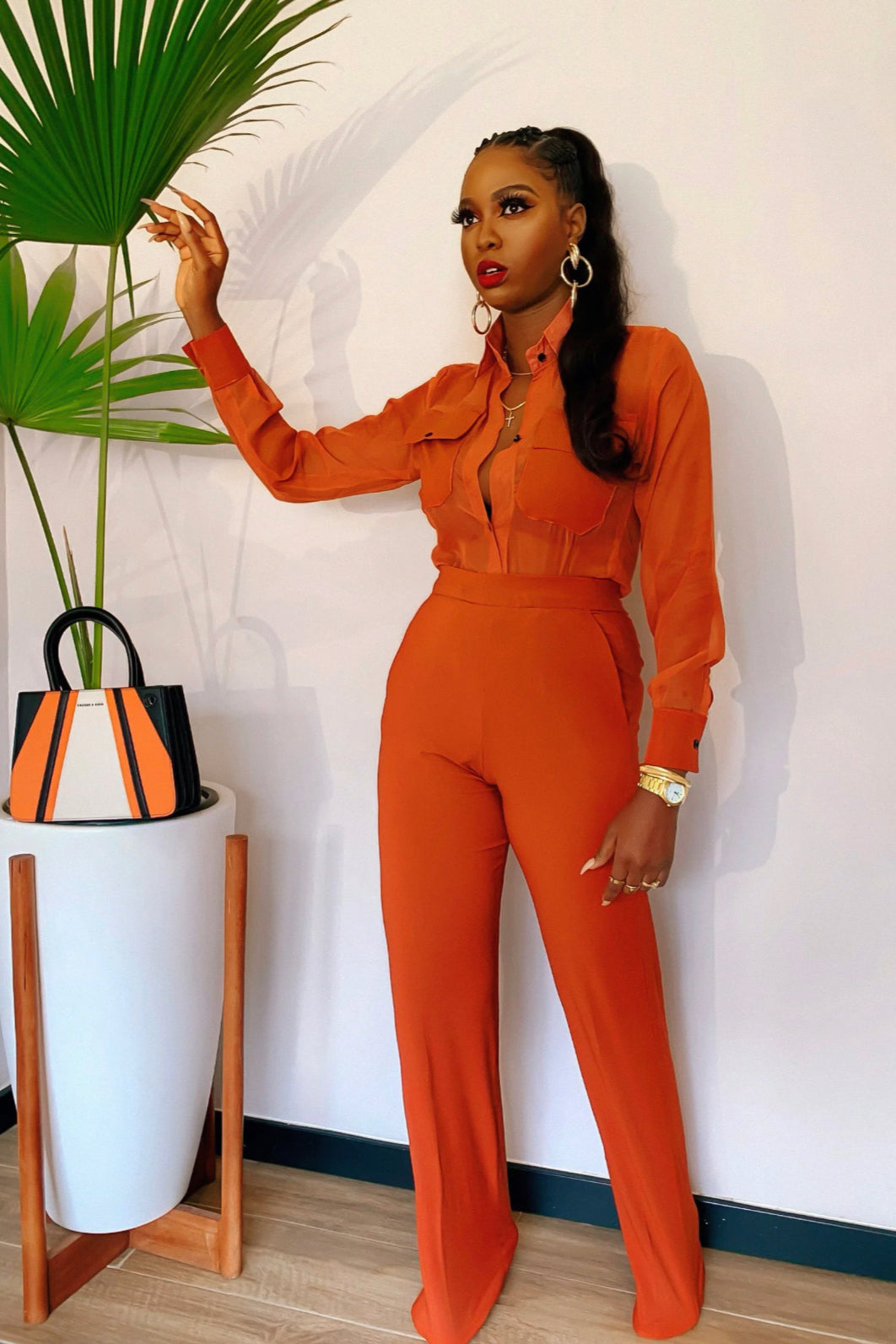 A model wearing an orange long-sleeved chiffon shirt with button closures and double flap pockets, paired with high-waisted pants featuring pockets on both sides, side zip and metal hook closure.