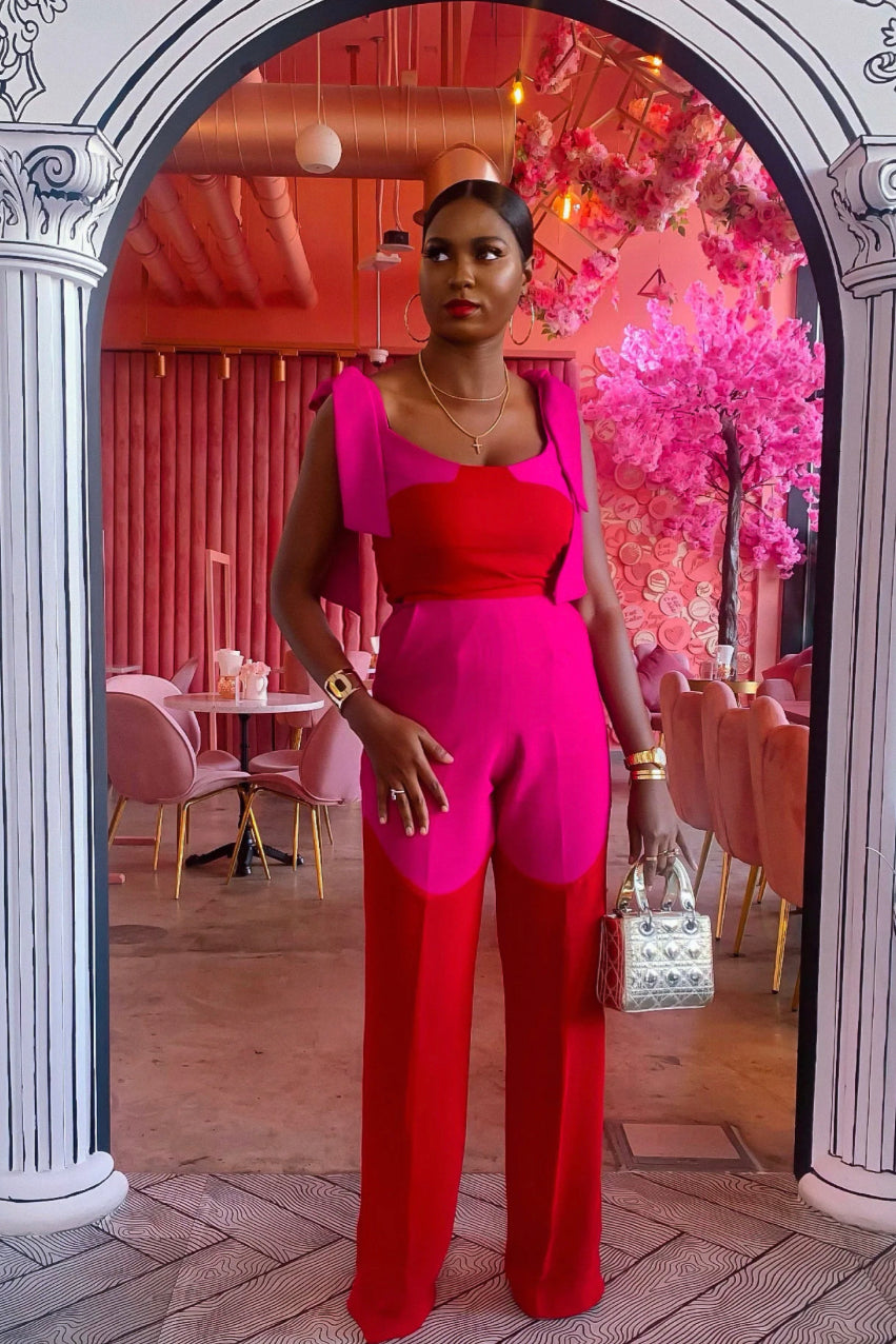 A person wearing a pink tie crop top and high-waist pants with a side zip and metal hook closure, styled with a Dior silver mini bag. Model is standing in a doorway with a reflective pink background. 