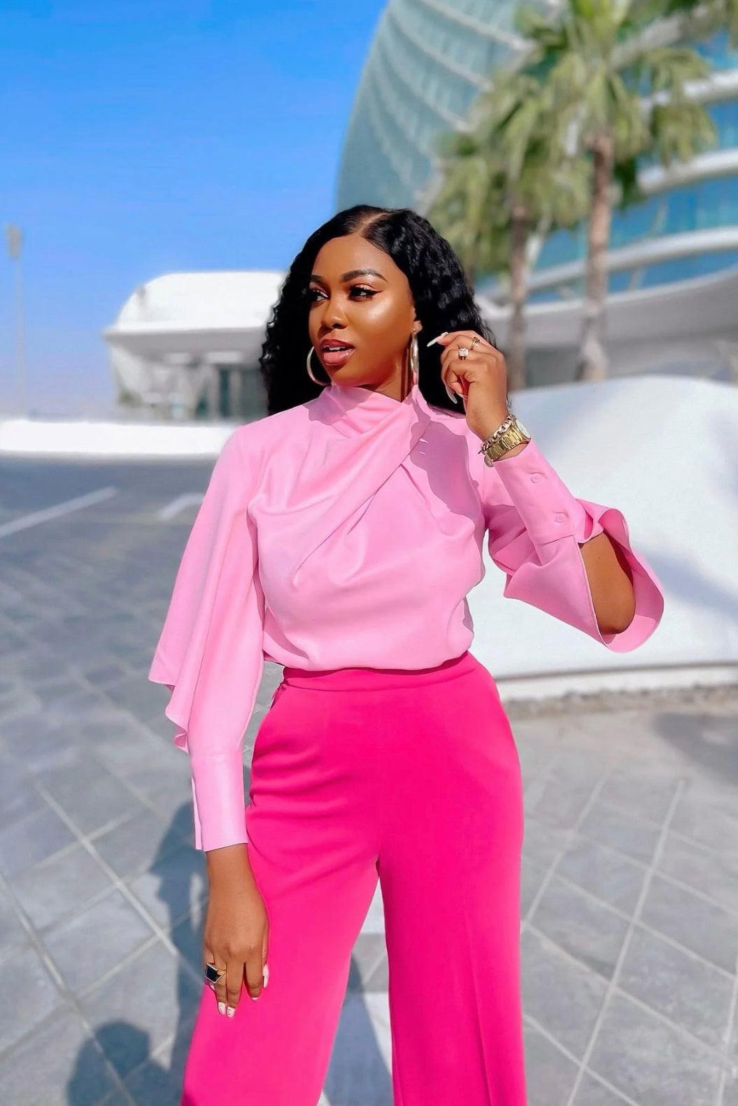 A female model wearing a pink blouse with a draped high neck and long sleeves with slits on the cuffs, paired with matching high-waisted pink pants. She is standing outdoors in Dubai with buildings in the background.