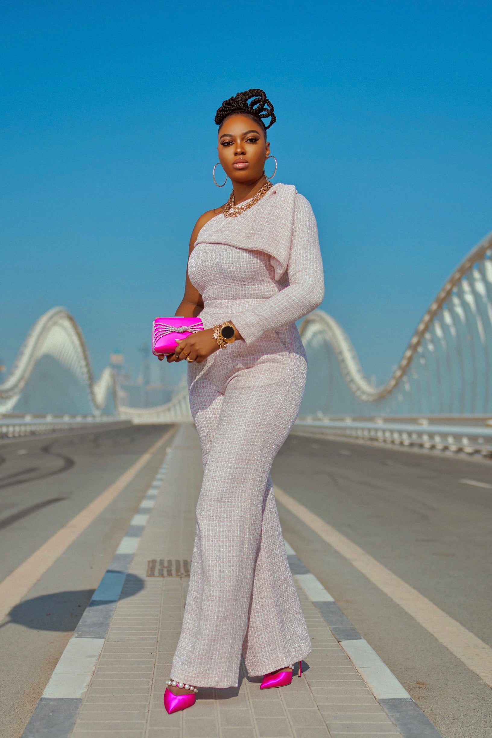 Model stands on Dubai’s Meydan Bridge in a luxury pink tweed two-piece set—one-shoulder long-sleeve bow-detail top with a gold shoulder button, paired with high-waist pants, styled with a hot pink Jimmy Choo clutch and heels.