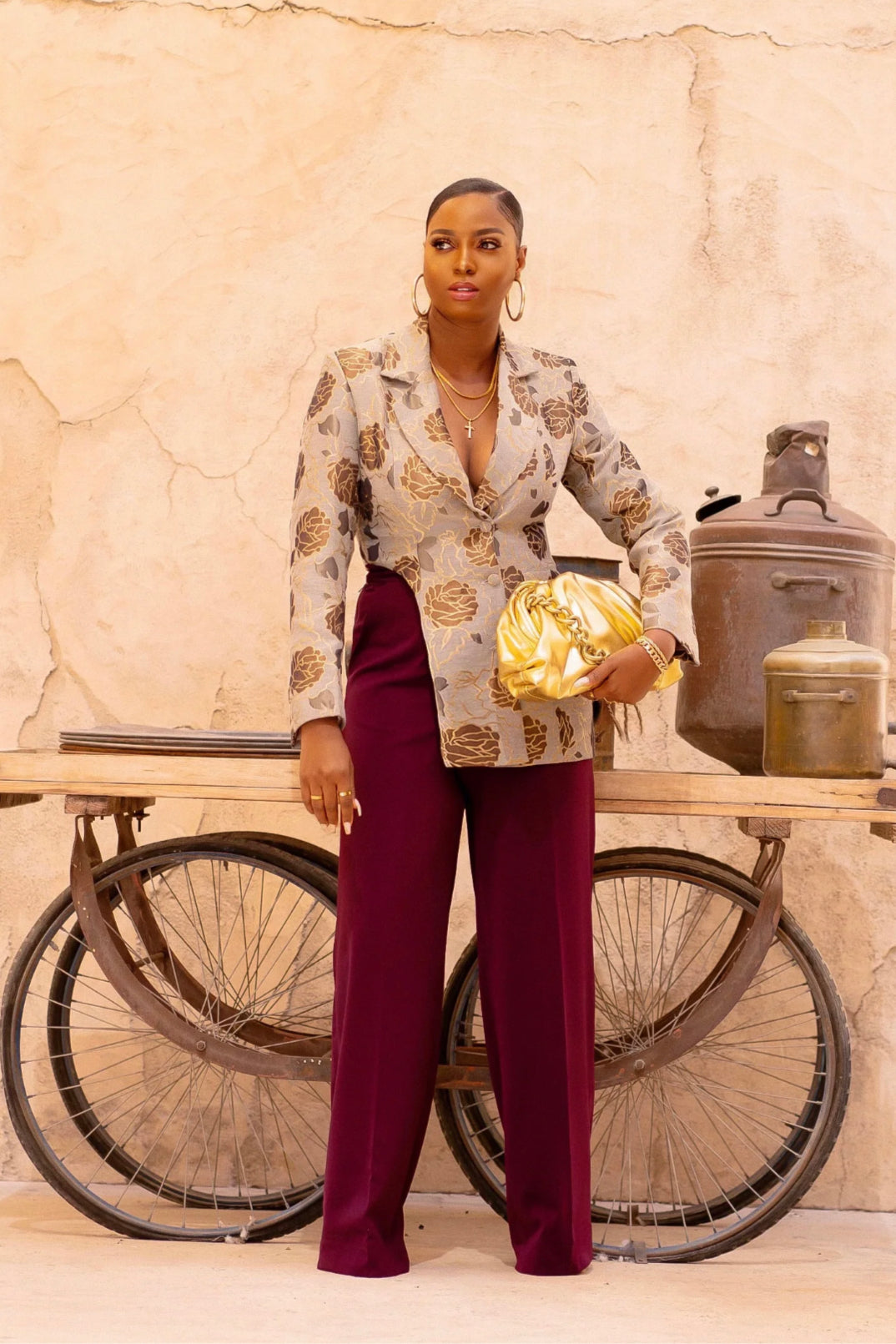 A tall model in a grey and gold floral brocade blazer with a signature side cut, styled with burgundy trousers and gold accessories.