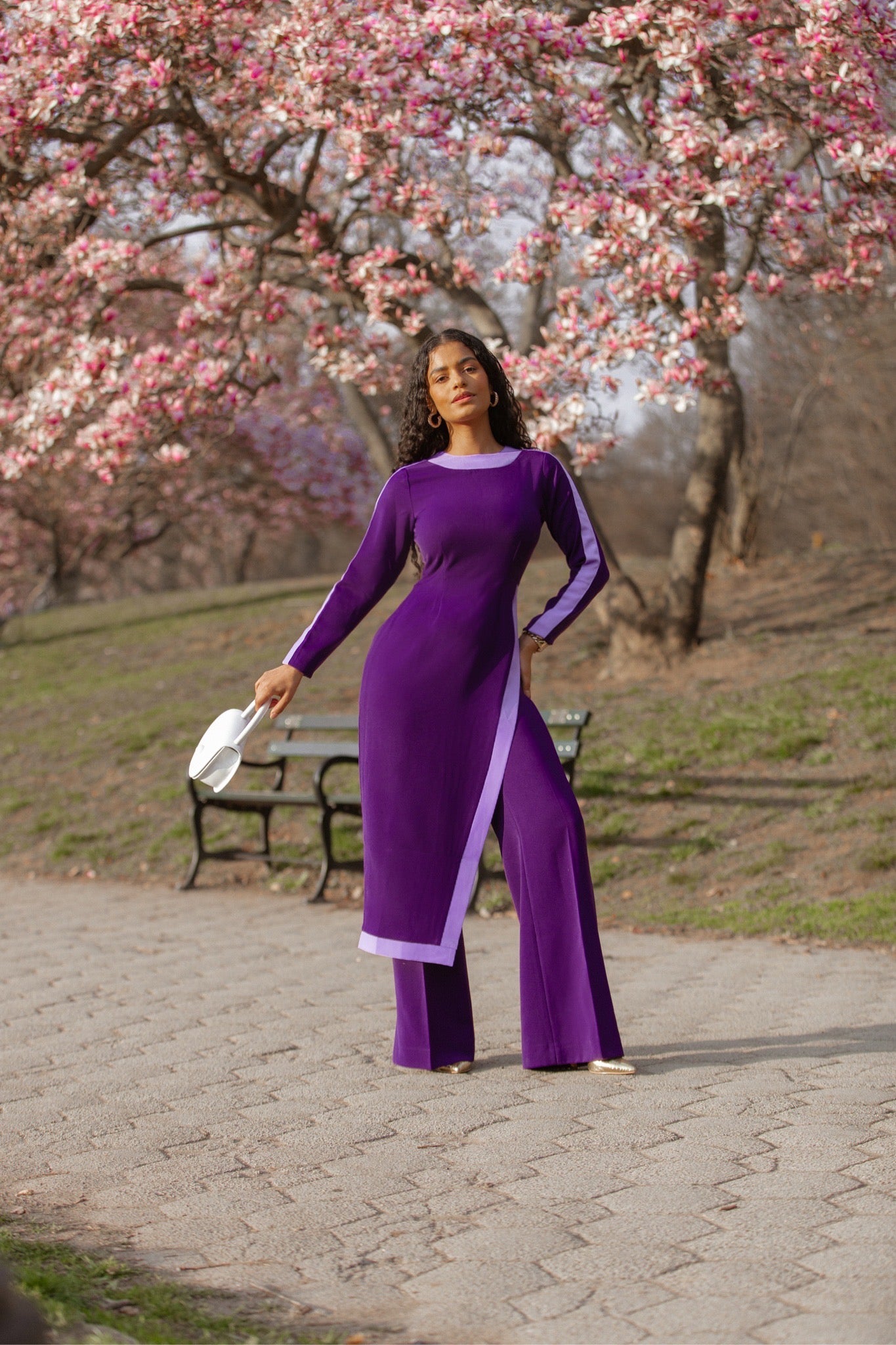 Model wearing purple and lilac color-block tunic with side slit over matching straight-leg pants in a spring park setting.
