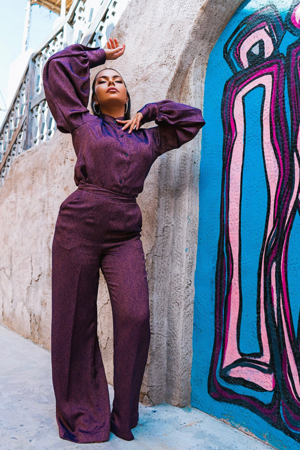 A Middle-eastern Model posing against a colorful graffiti background in Dubai, wearing a purple sleeveless Sabrina set with exaggerated balloon sleeves and a bishop collar neck.