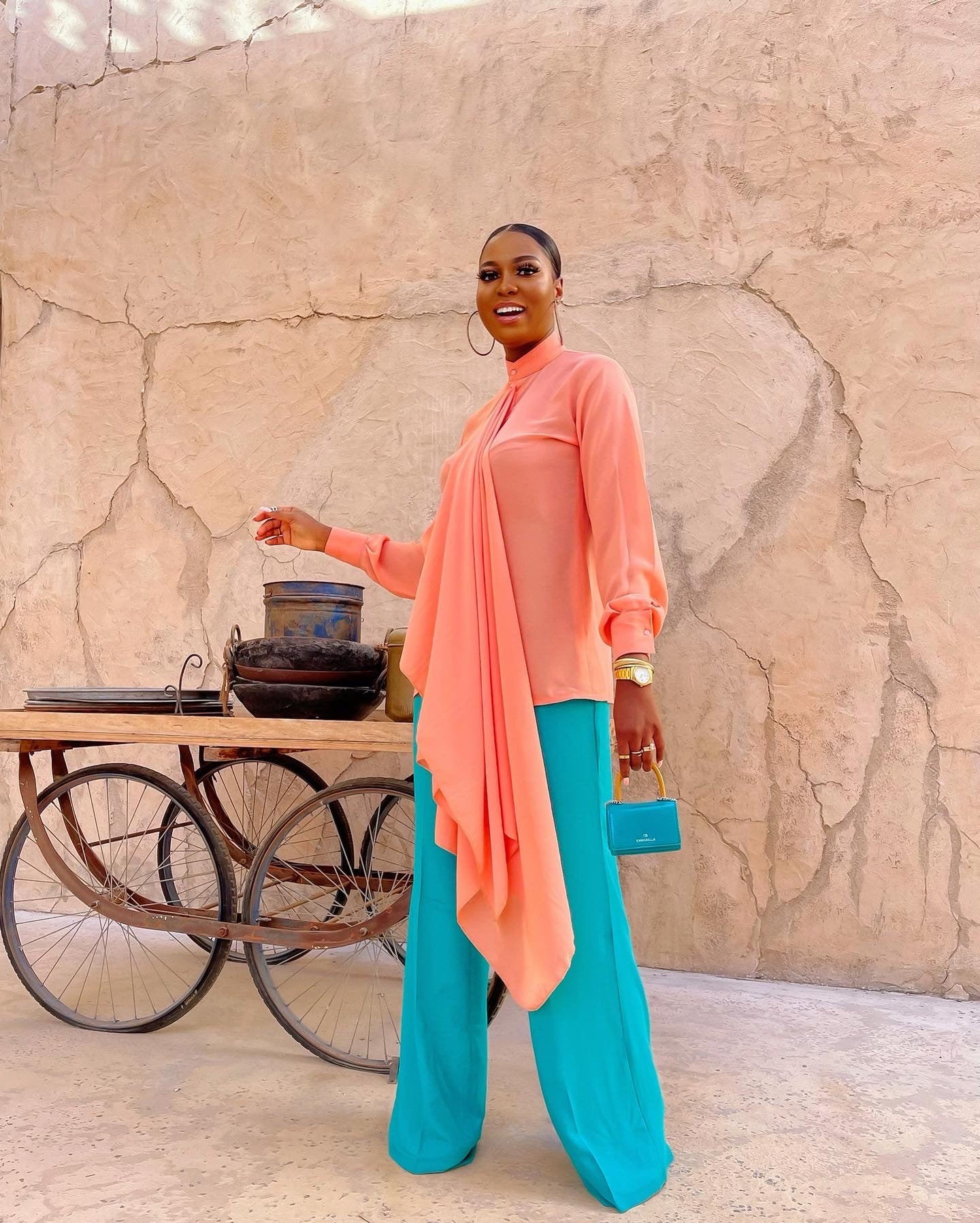 Model wearing Tabitha Set featuring peach bishop collar blouse tucked out, and featuring a long neck drape and teal high-waist straight trouser, holding a teal mini bag. 