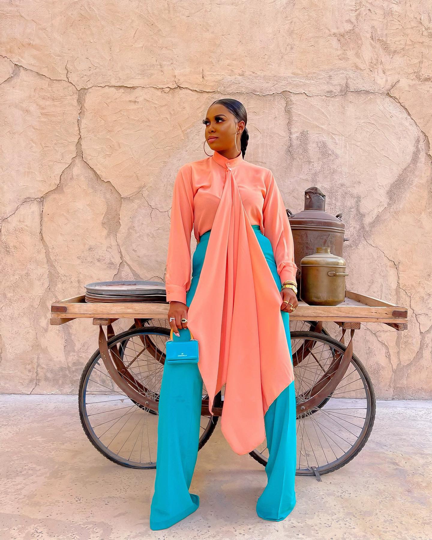 Model wearing an elegant Olarsgrace Tabitha Set featuring peach bishop collar blouse with long neck drape and teal high-waist wide-leg pants, holding a teal mini bag for a chic look.